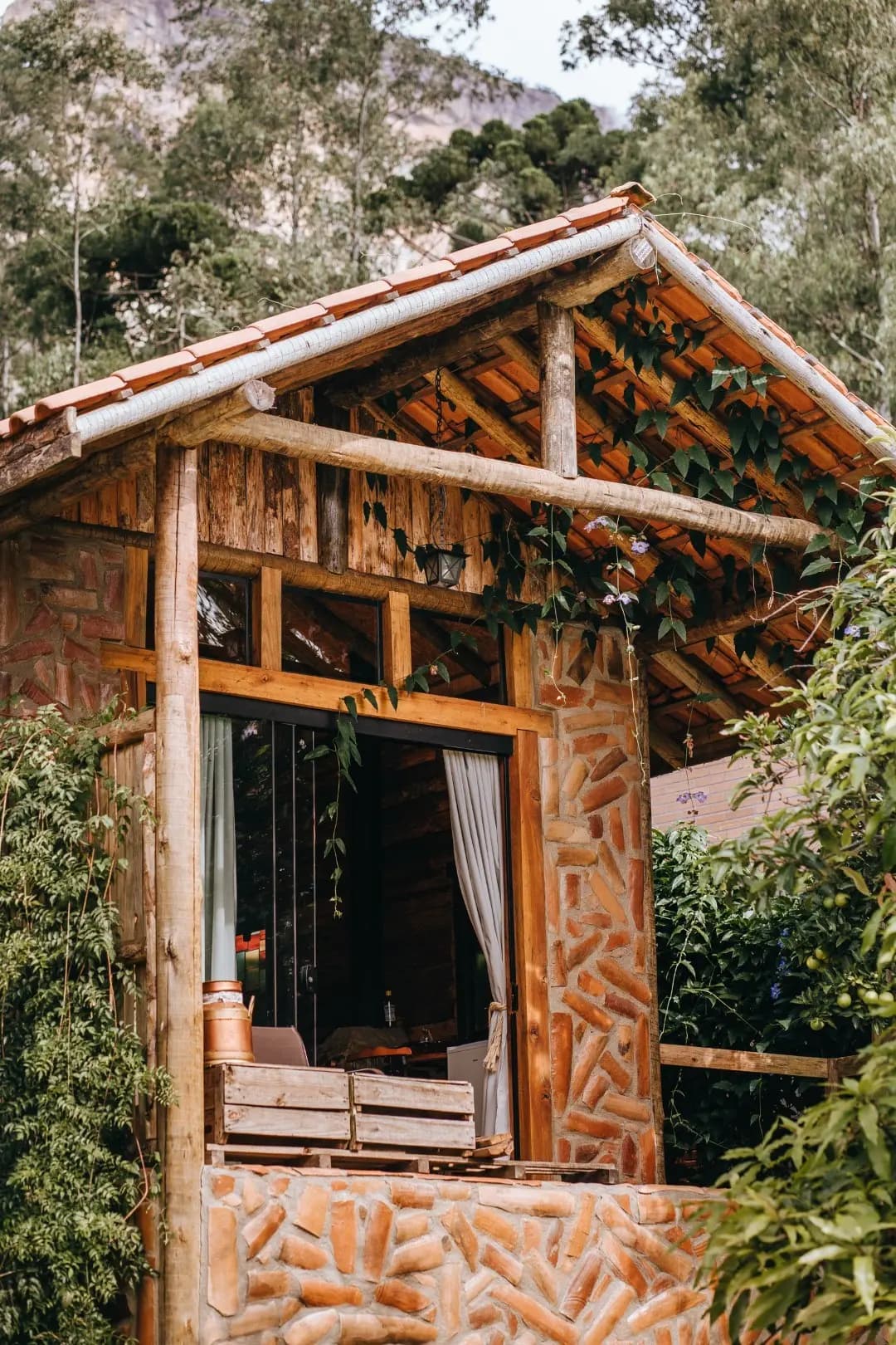 Fachada da Cabana Ametista em madeira, pedra e tijolo, com porta de vidro para a varanda e trepadeiras no telhado
