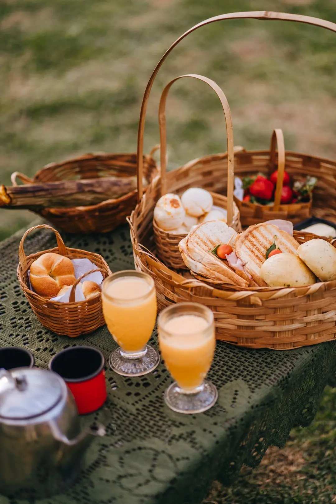 Café da manhã servido no gramado: cestas de pães, pão de queijo, sanduíches, sucos e canecas esmaltadas