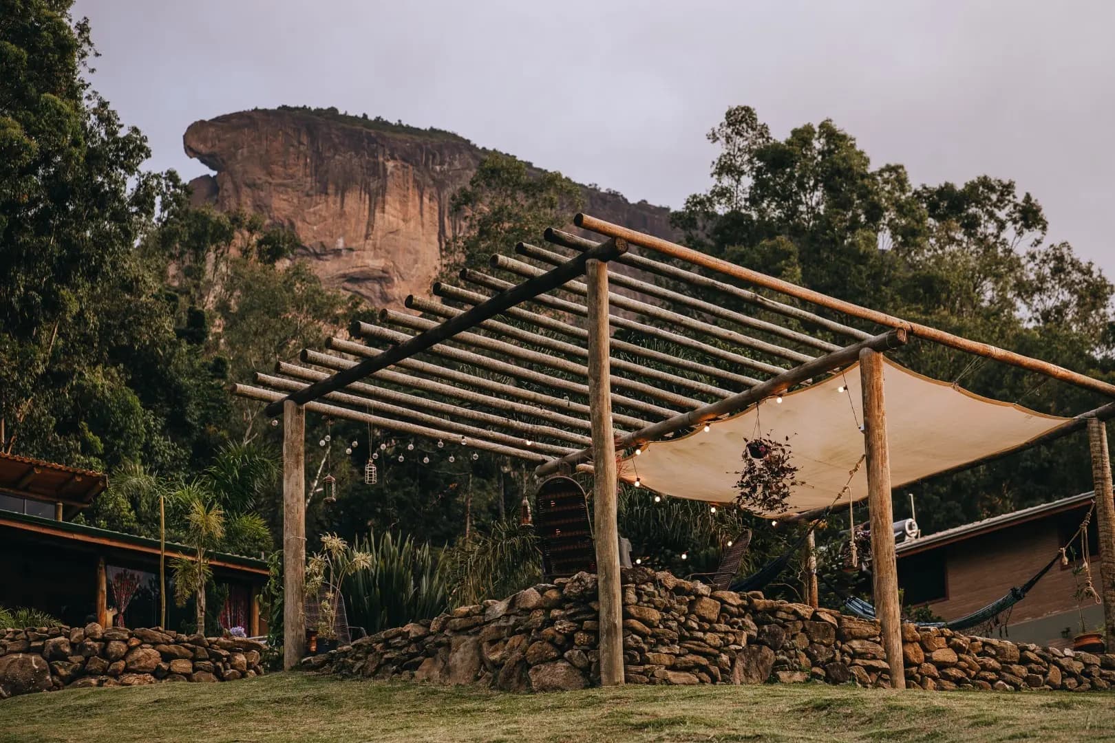 Pergolado de troncos com toldo de lona e redes, tendo a Pedra do Baú ao fundo