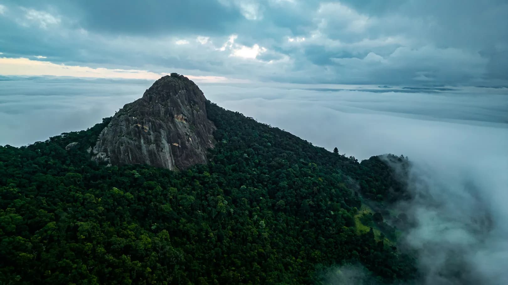 Monólito do complexo do Baú visto do alto ao amanhecer, cercado de mata e acima de um mar de nuvens
