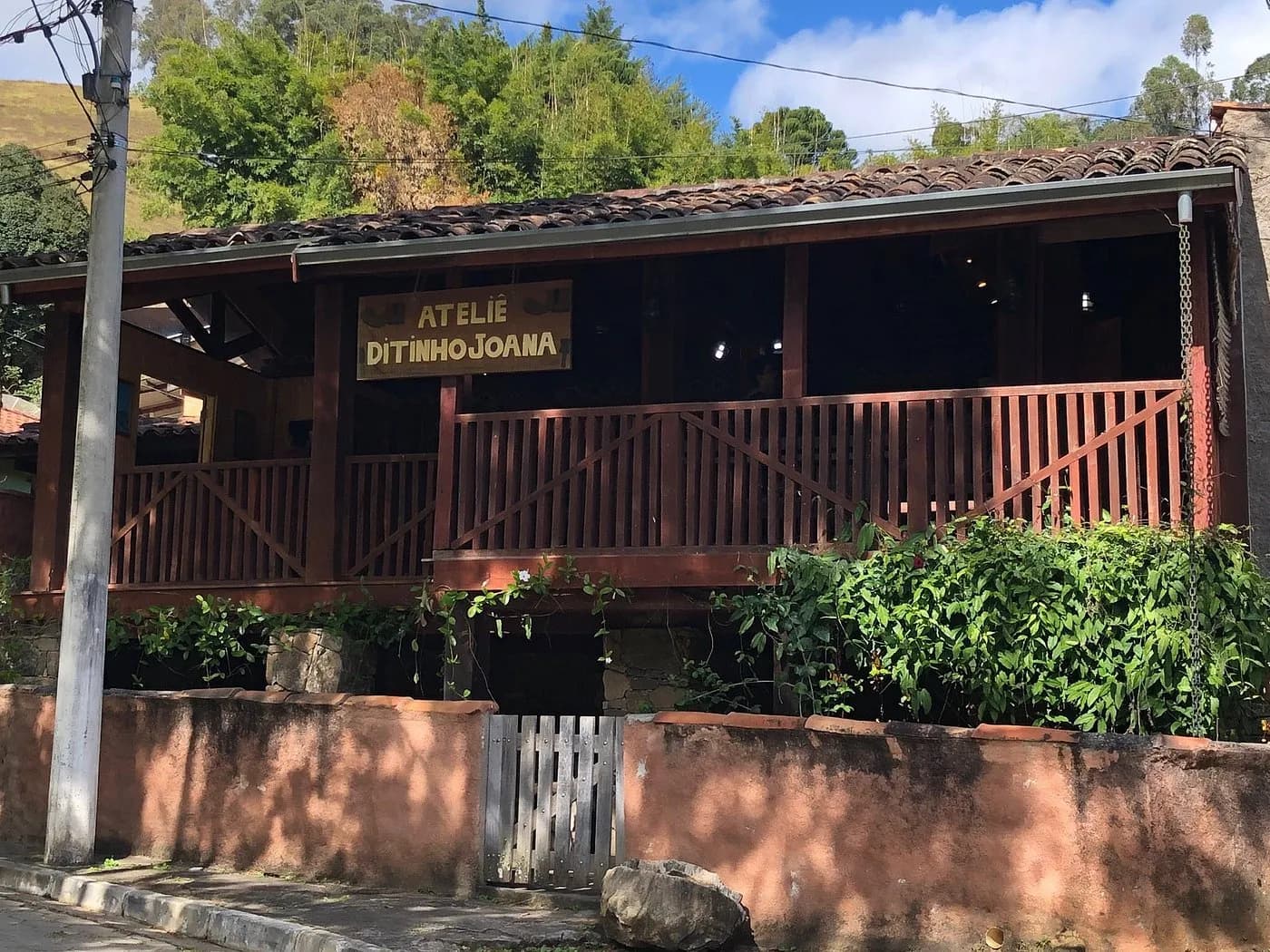 Fachada do Ateliê Ditinho Joana, casa de madeira avermelhada com varanda, placa entalhada e muro de tijolo