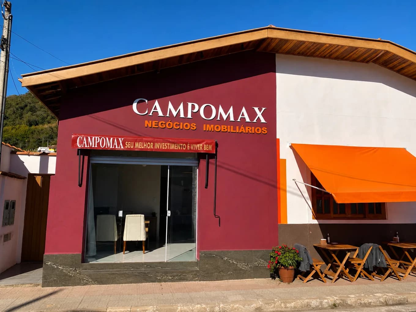 Fachada vinho da CAMPOMAX Negócios Imobiliários em dia de céu azul, com o letreiro branco e dourado sobre a vitrine de vidro