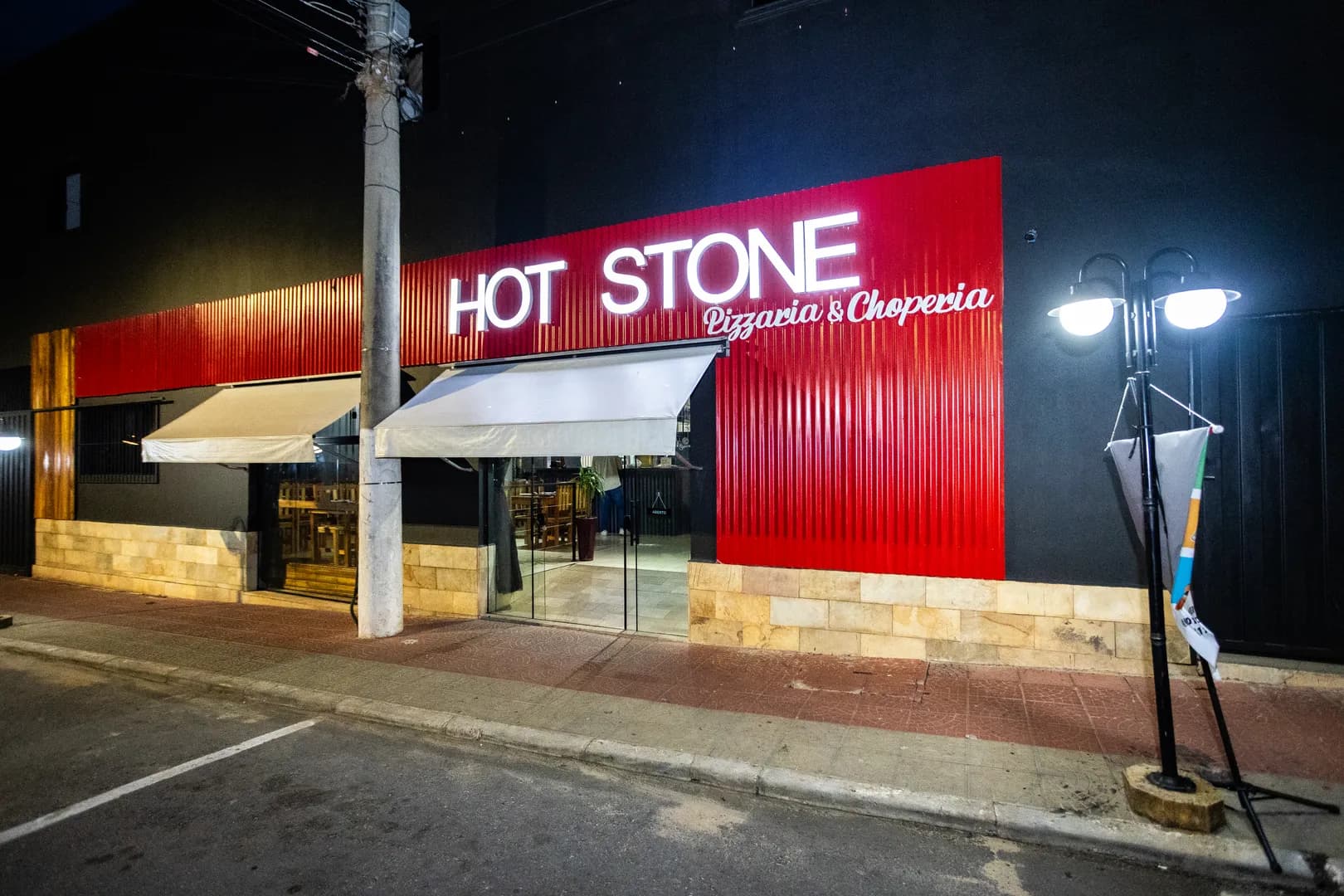 Fachada da Hot Stone à noite, com o letreiro branco iluminado sobre painel vermelho canelado e os toldos claros da entrada