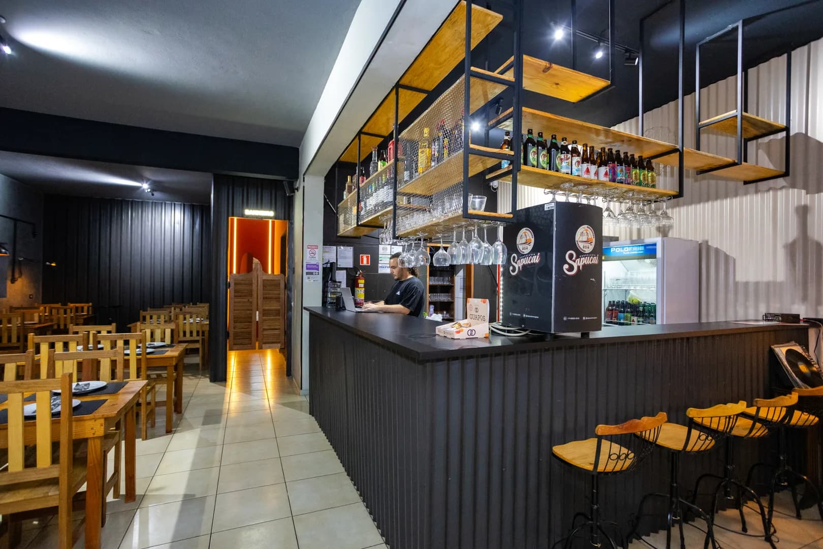 Balcão preto da Hot Stone visto do corredor do salão, com prateleiras suspensas de garrafas, taças penduradas, geladeira de bebidas e a coluna de chope da Cervejaria Sapucaí