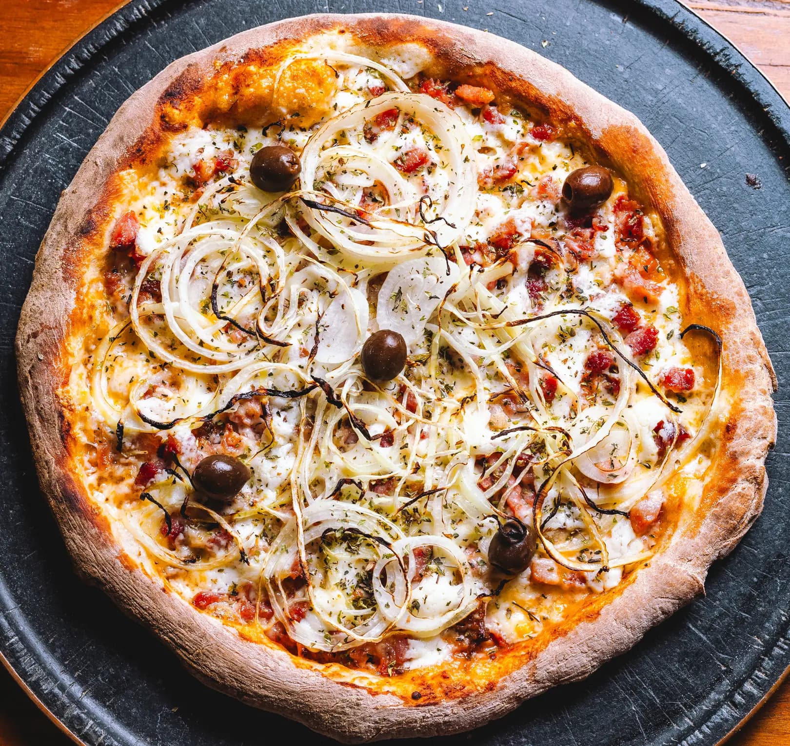 Pizza inteira na chapa preta, com muçarela, calabresa, rodelas de cebola douradas, azeitonas e orégano, e a borda alta