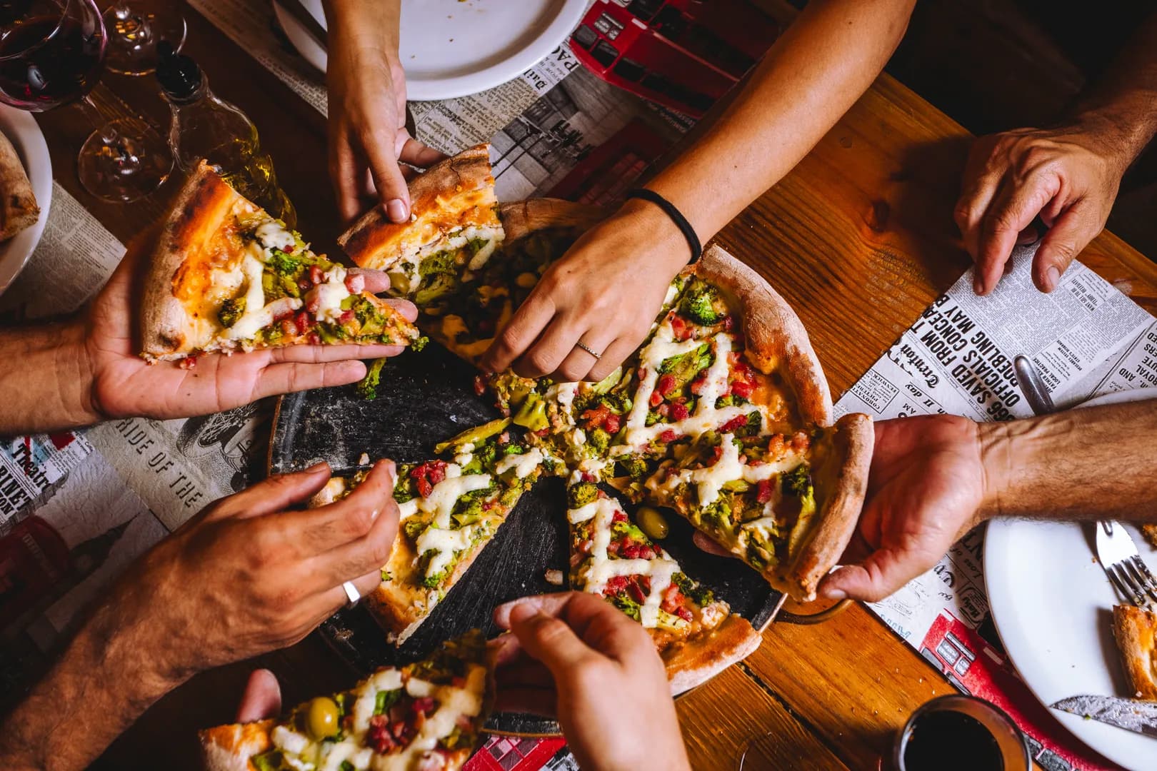Seis mãos tirando fatias ao mesmo tempo de uma pizza de brócolis com calabresa e azeitona, na chapa preta sobre a mesa de madeira forrada de jornal