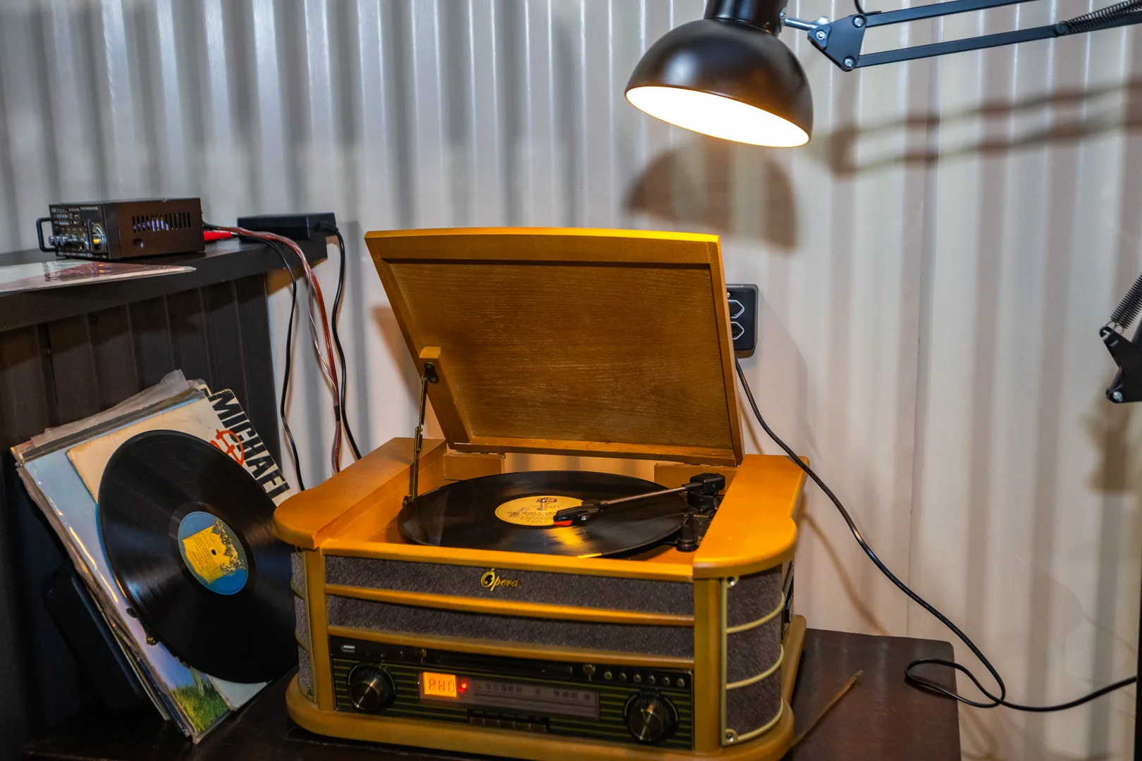 Vitrola de madeira clara tocando um disco no salão da Hot Stone, com a pilha de LPs encostada ao lado e a luminária articulada acesa por cima