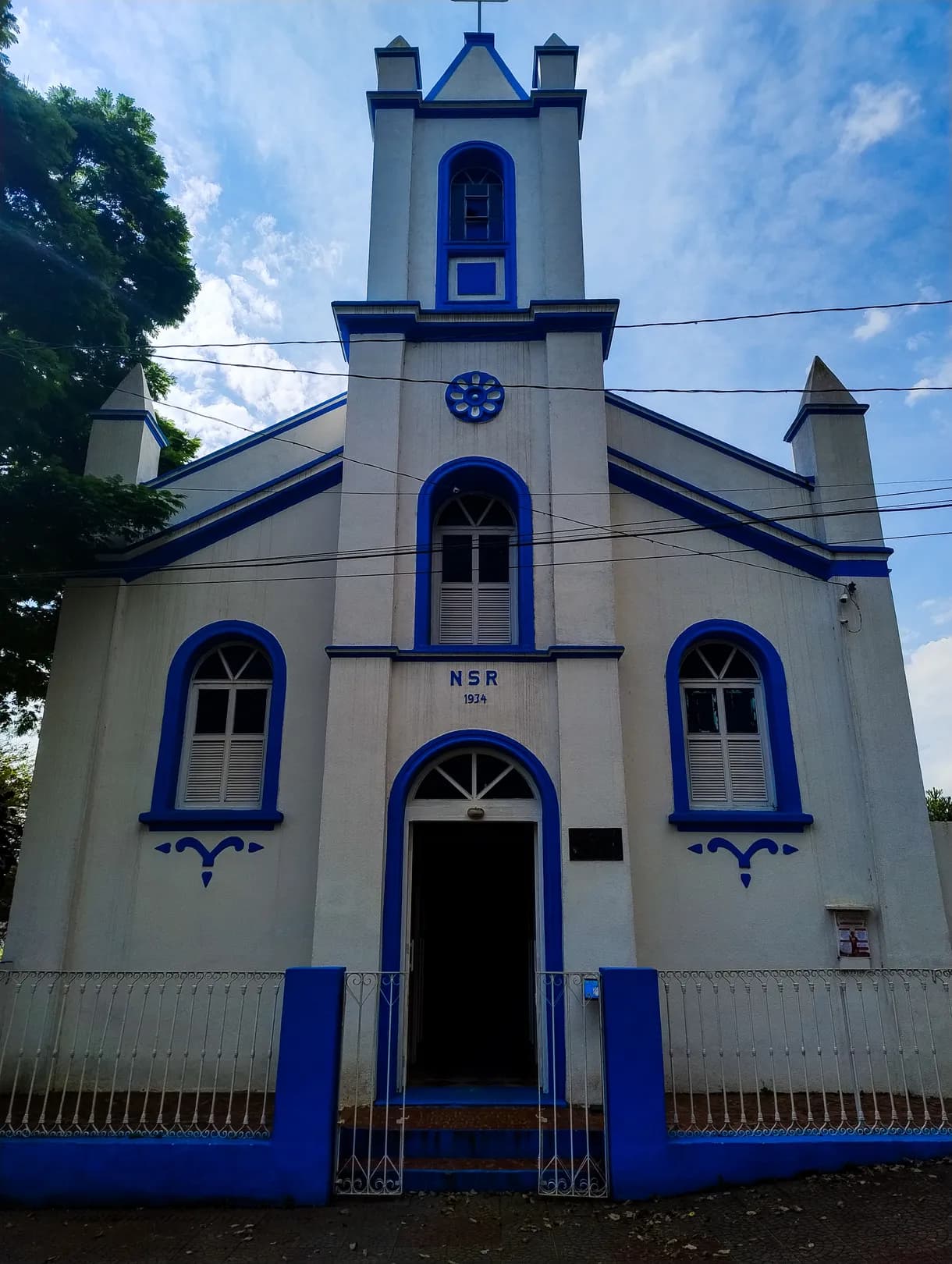 Fachada branca e azul da Igreja Nossa Senhora do Rosário, com torre única, rosácea e gradil azul na frente
