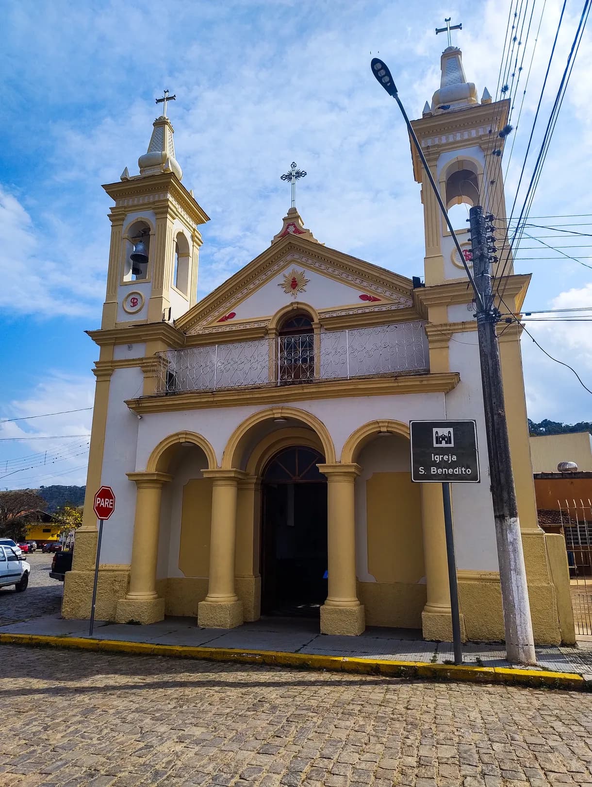 Fachada ocre e branca da Igreja São Benedito, com dois campanários e três arcos na entrada, na rua de paralelepípedo
