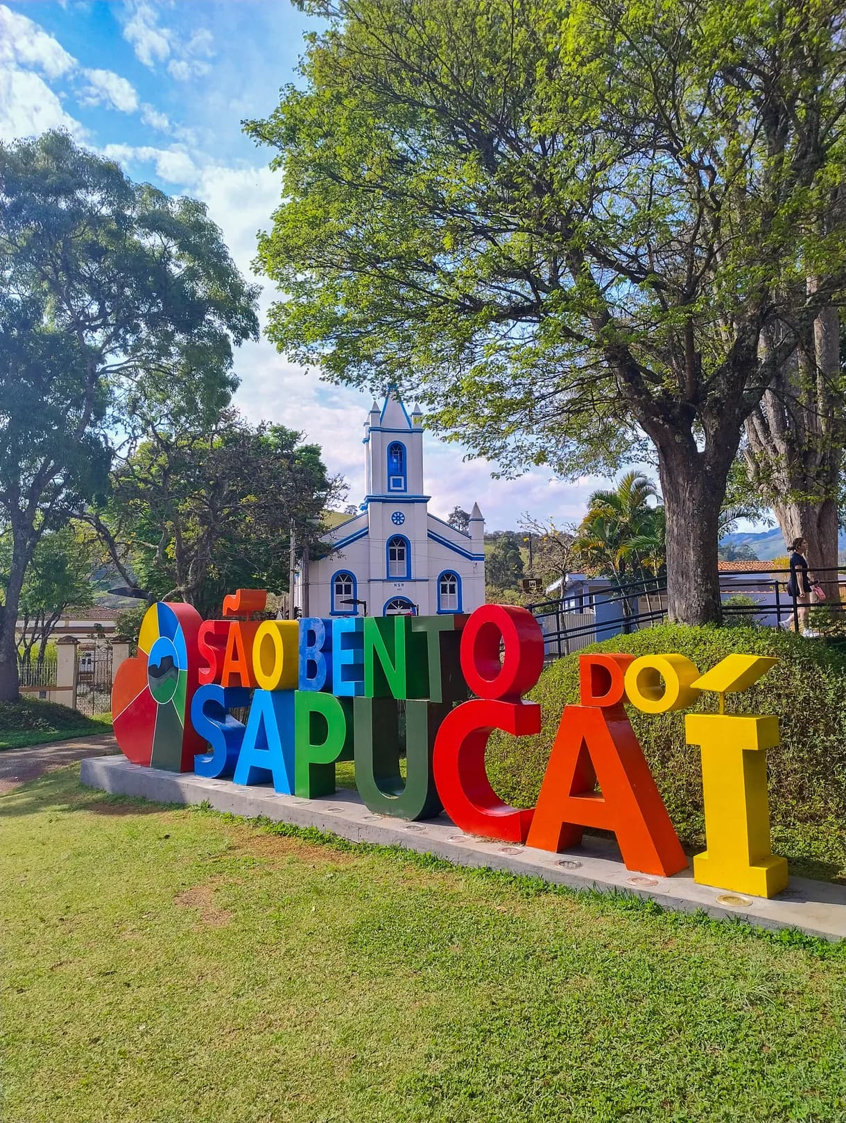 Letreiro colorido de São Bento do Sapucaí no gramado da praça, com a igreja azul e branca logo atrás
