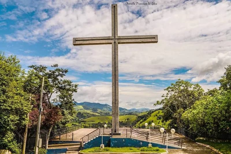 Cruz de cimento do Mirante do Cruzeiro sobre a plataforma de piso claro, com escadas, guarda-corpo e os morros verdes ao fundo