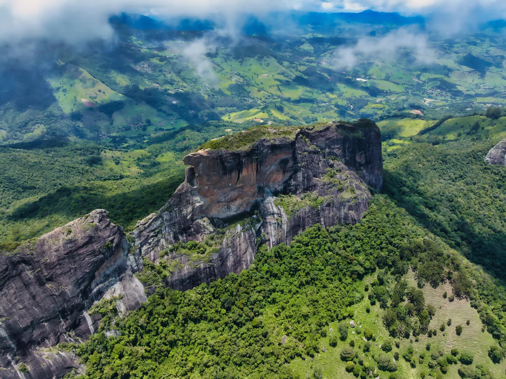 Vista aérea do complexo do Baú entre nuvens baixas, com o paredão de rocha cercado de mata