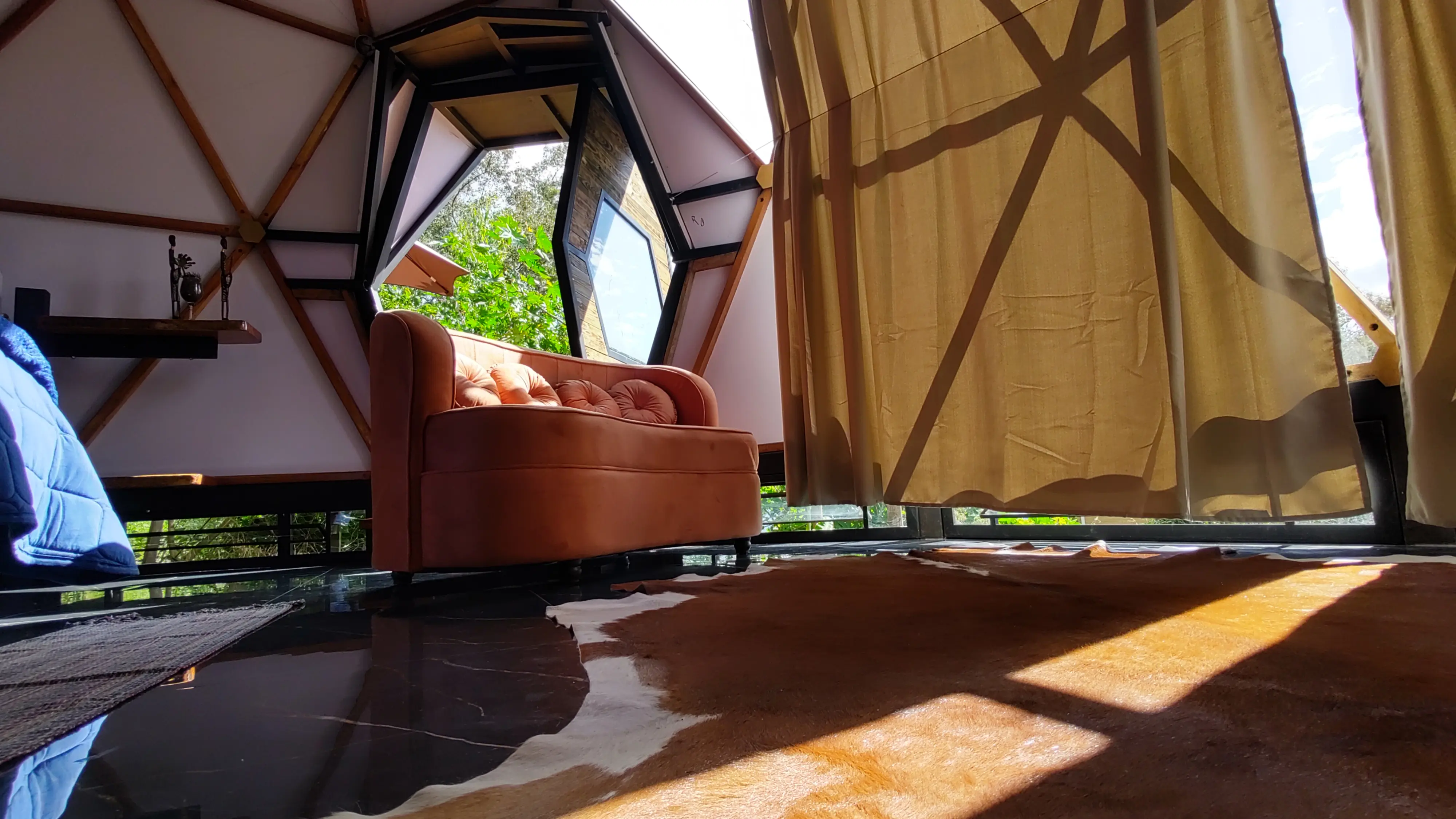 Interior do domo Colmeia: sofá de veludo salmão junto à janela hexagonal, tapete de couro e piso escuro