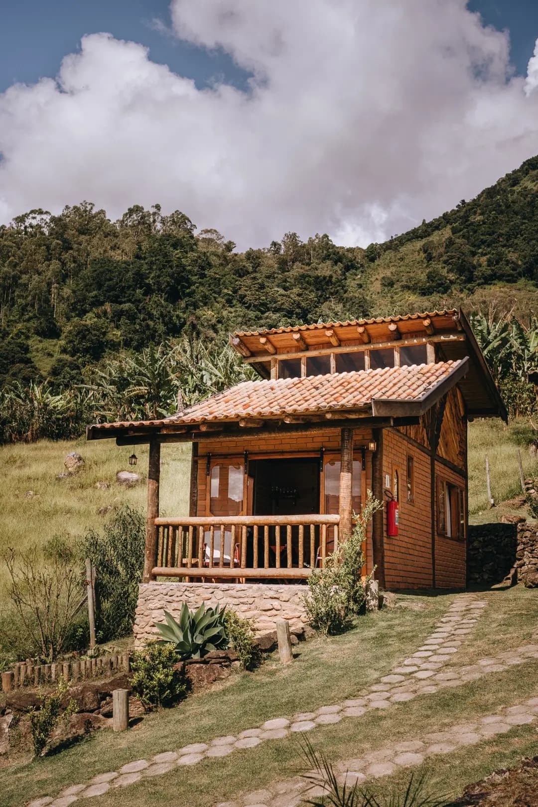 Chalé Turmalina visto de fora, com telhado de telha e clarabóia, varanda de troncos, base de pedra e caminho de pedras no gramado