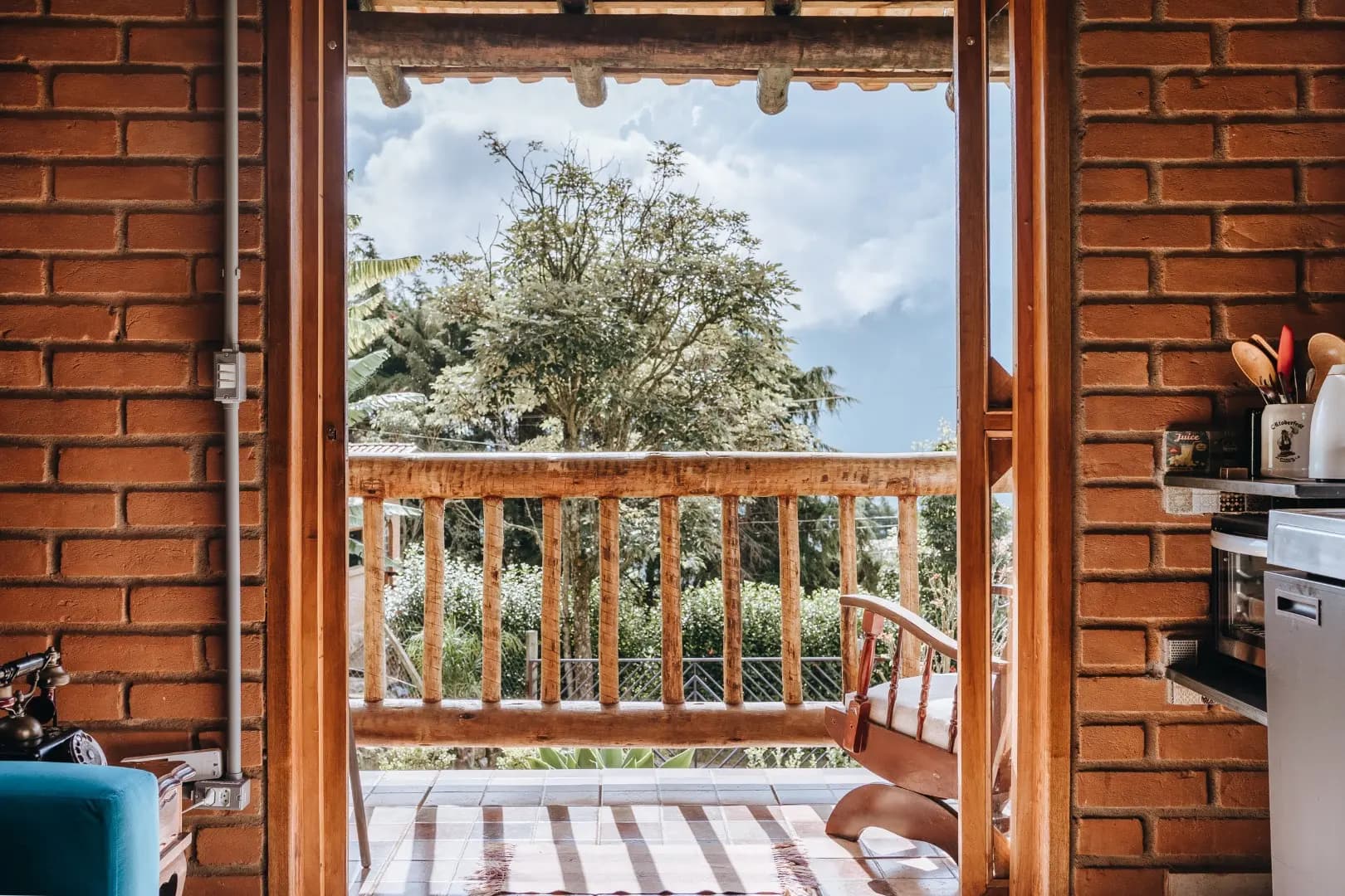 Vista da sala do Chalé Turmalina para a varanda, com porta de vidro aberta, parede de tijolo aparente e forno na bancada